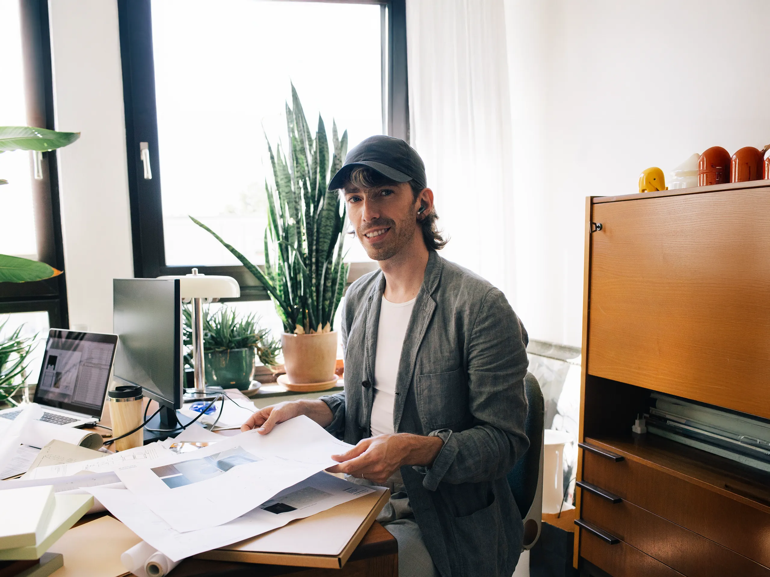 Man with baseball cap working at a desk with many plants.