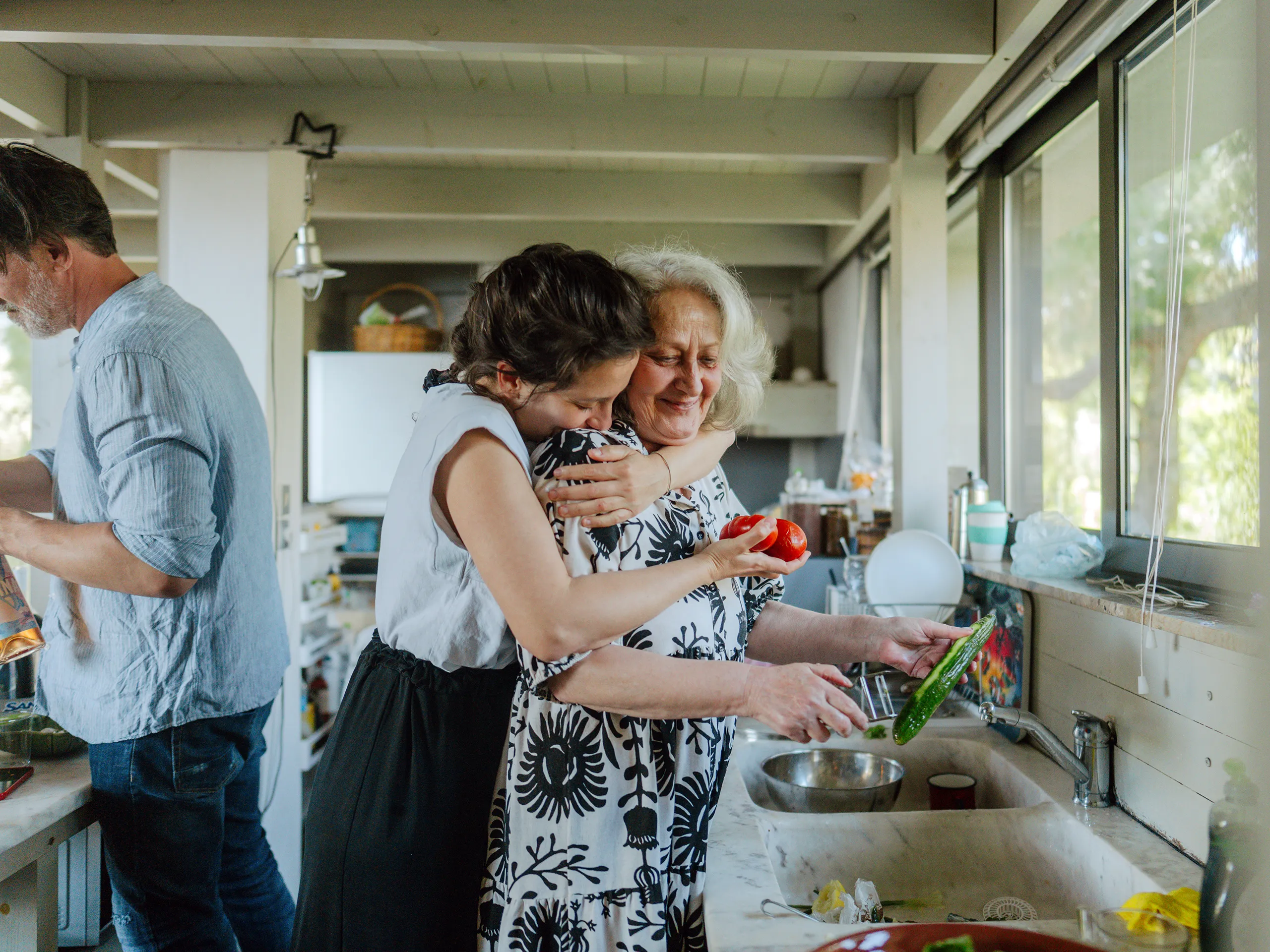 Older woman cooks in the kitchen while a younger woman hugs her.