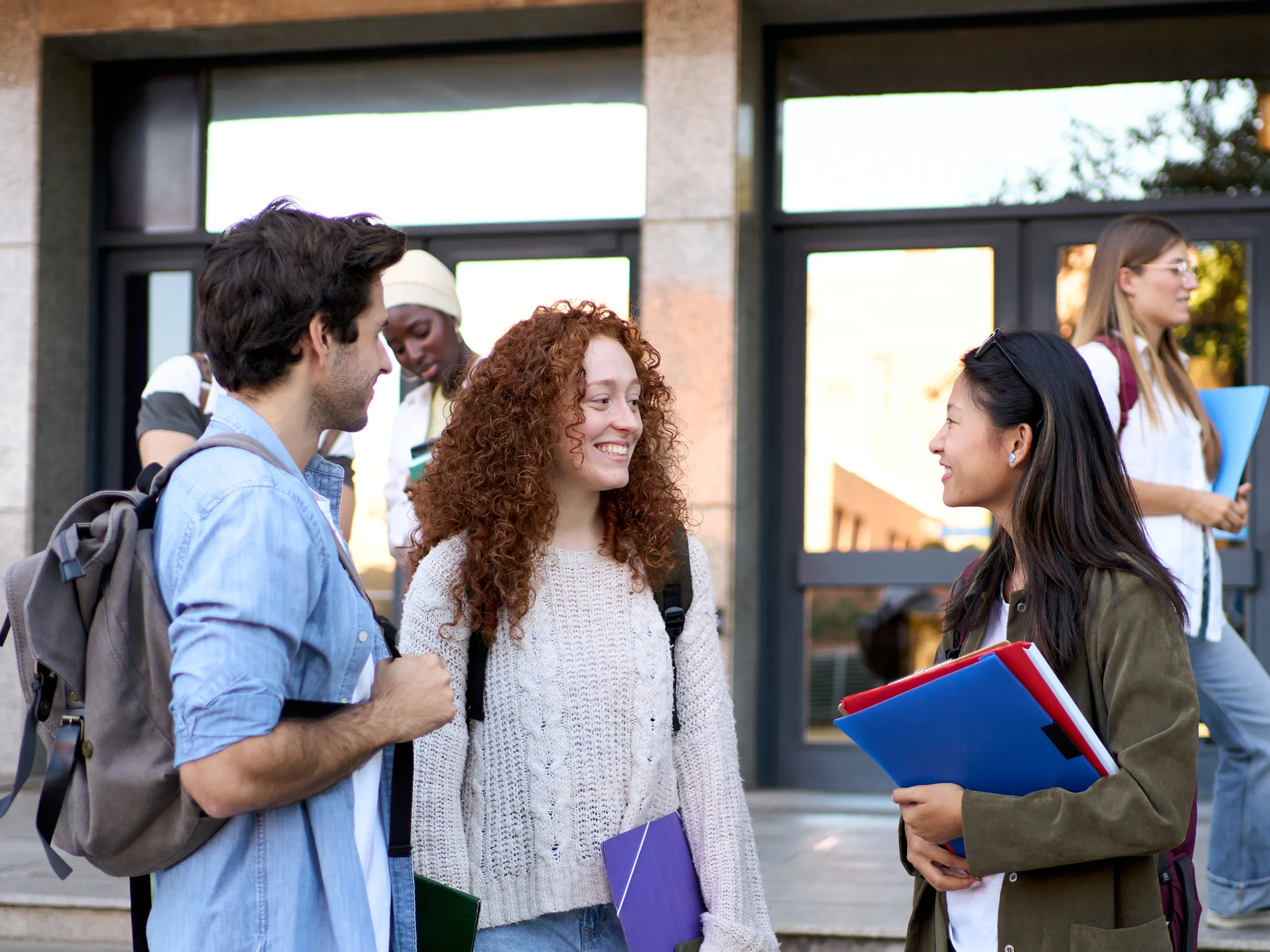 Three young people are smiling and talking in front of a campus building.
