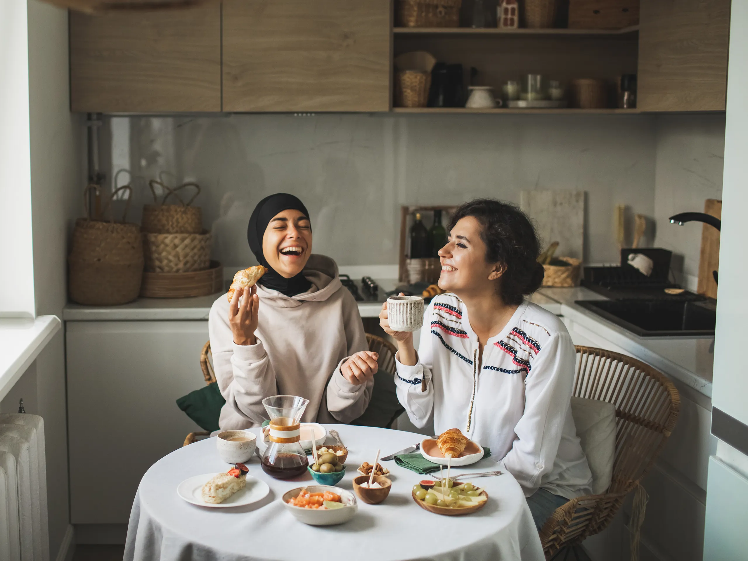 Two women laugh while having breakfast at a set table in the kitchen.